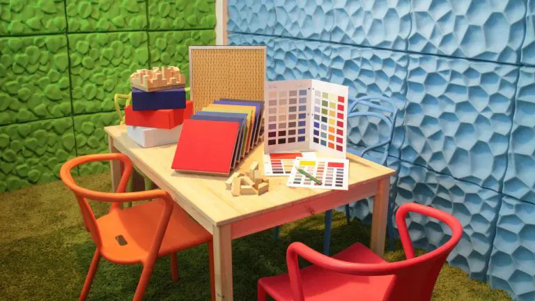 A small wooden table with colorful chairs, color swatches, books, and wooden puzzle blocks, set in a room with textured green and blue walls, green carpet, and subtle acoustic treatment for a cozy atmosphere.