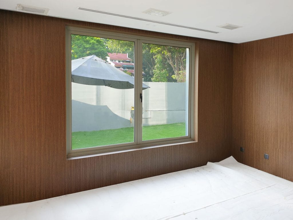 A room with Timberix Residential Acoustic Solutions features brown wooden walls and a large window overlooking a green lawn, gray fence, and trees. An outdoor umbrella is visible, while the floor inside is covered with a white sheet.