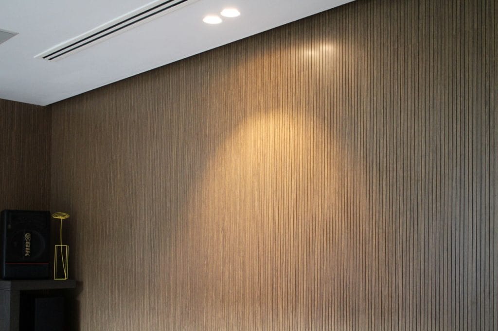 A brown, vertically striped Timberix Residential Acoustic Solutions wall is illuminated by two ceiling spotlights, creating a bright oval-shaped patch. In the corner, a black speaker rests on a dark shelf with a small yellow wireframe decoration.