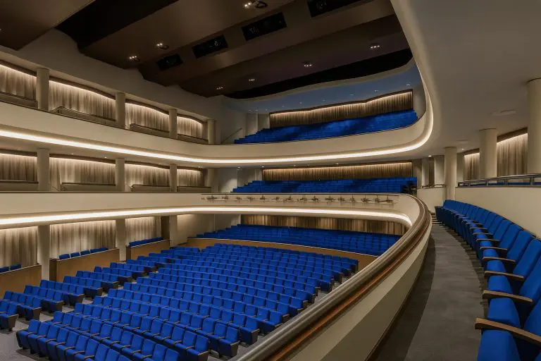 A modern, spacious theater auditorium with multiple balconies and rows of empty blue seats, illuminated by soft, warm lighting. Designed with curved lines and neutral-colored walls, the space boasts excellent theater acoustics for an immersive experience.