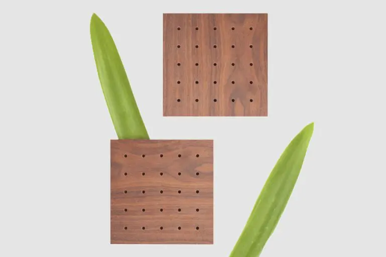 Two square wooden coasters, inspired by timber acoustic panels, feature small holes in a grid pattern. They are arranged diagonally with two green leaves behind them on a plain light background.