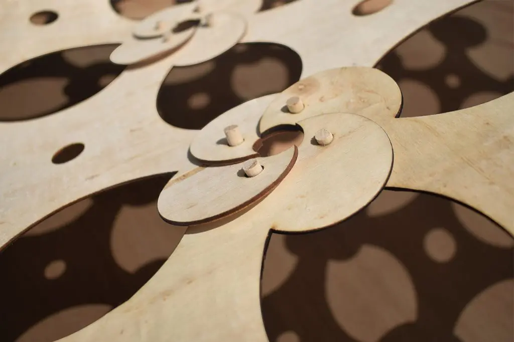 Close-up view of an intricate wooden structure with interlocking, rounded shapes and cut-out patterns, resembling timber acoustic panels, showing pegs connecting several overlapping pieces.