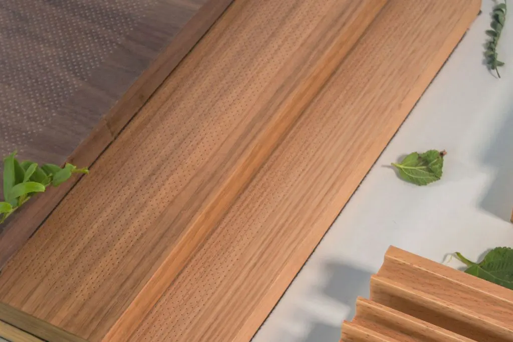 Close-up of micro perforated wood panels with finely drilled holes, arranged on a light surface and accented with a few green leaves. The panels display varied wood tones and distinct grain patterns.