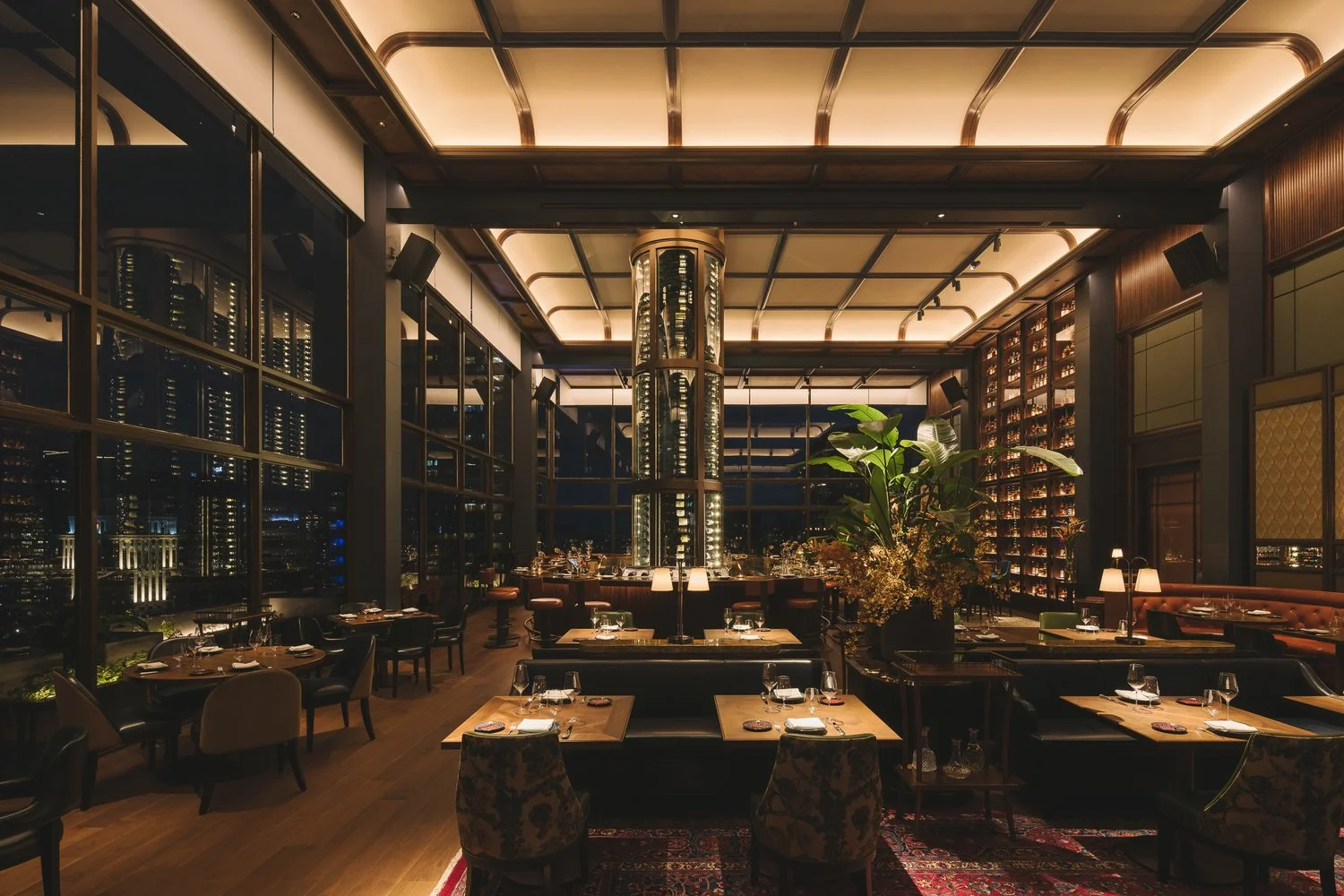 A spacious, elegant restaurant with floor-to-ceiling windows, warm lighting, plush seating, and stylish decor. Enhanced by Timberix Acoustic Projects, guests dine comfortably while enjoying city lights through the windows at night.