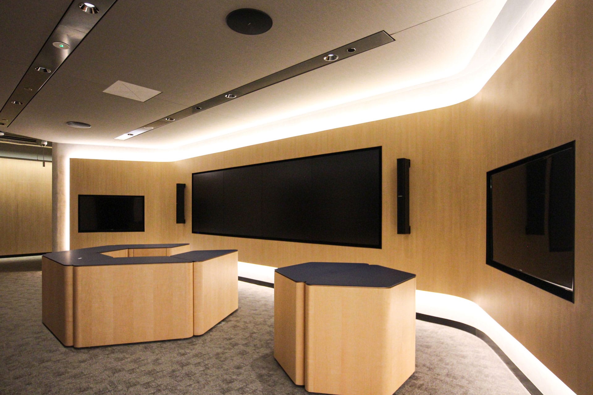 A modern, minimalist meeting room with wood-paneled walls featuring Timberix Office Acoustic Solutions, large flat-screen monitors, two polygonal tables, gray carpet, and recessed ceiling lighting.