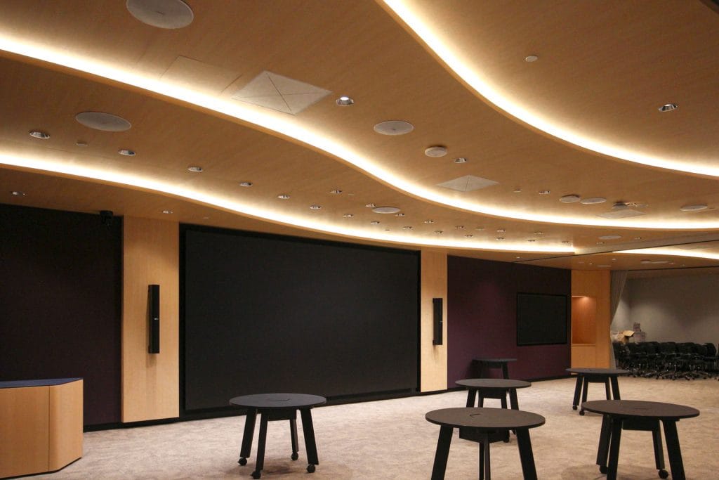 A modern conference room with a large blank screen, curving wooden ceiling featuring Timberix Office Acoustic Solutions and recessed lighting, plus several round black tables. The room has carpeted floors and empty chairs visible in the background.