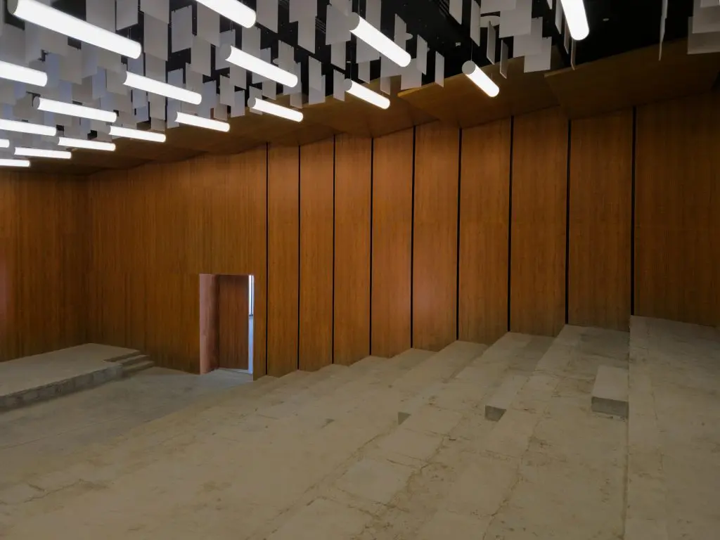 An empty amphitheater-style room with tiered stone seating, vertical wooden wall panels by Timberix Office Acoustic Solutions, a doorway with light shining through, and modern rectangular light fixtures with hanging ceiling panels.
