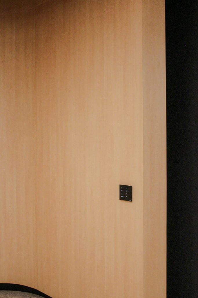 A light FSC® Acoustic Wood Panel-paneled corner wall with a small black control panel mounted on it; a narrow strip of black wall is visible on the right edge, highlighting traceability in design.