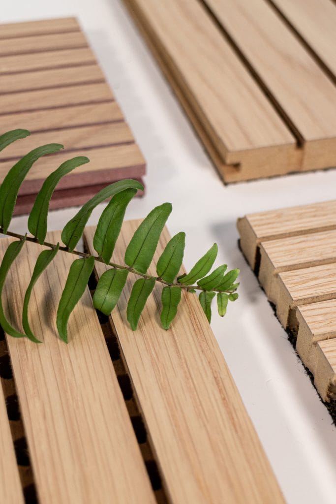 A green fern leaf lies across several rectangular panels of fire-safe timber cladding that doesn’t sacrifice craft, each with parallel grooves, arranged on a white surface.