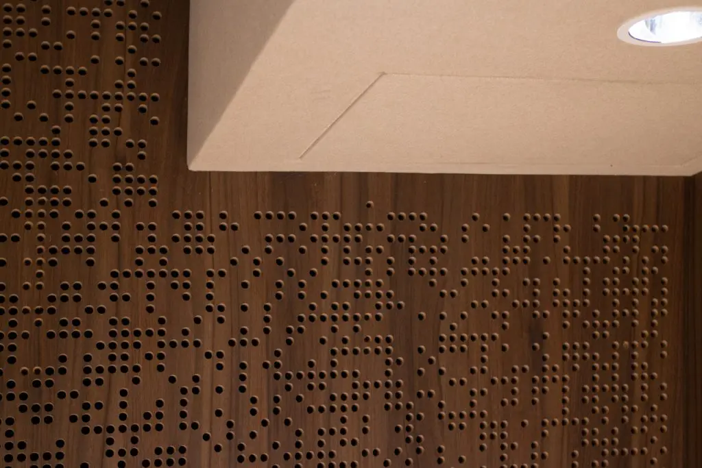 A Timberix Acoustic Projects wooden wall panel features a pattern of small round holes, beneath a beige acoustic ceiling panel with recessed lighting. The scattered holes in varying sizes create an abstract design.