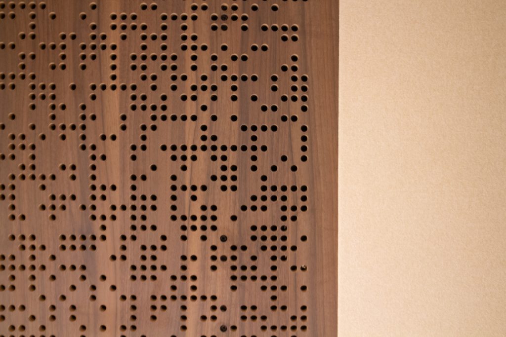 A close-up of a Timberix Acoustic Projects wooden panel with randomly spaced round holes on the left side, next to a plain beige wall on the right. The wood grain and perforations create a textured, modern design.