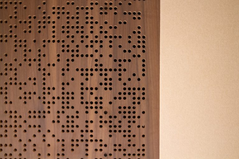 A close-up of a Timberix Acoustic Projects wooden panel with randomly spaced round holes on the left side, next to a plain beige wall on the right. The wood grain and perforations create a textured, modern design.