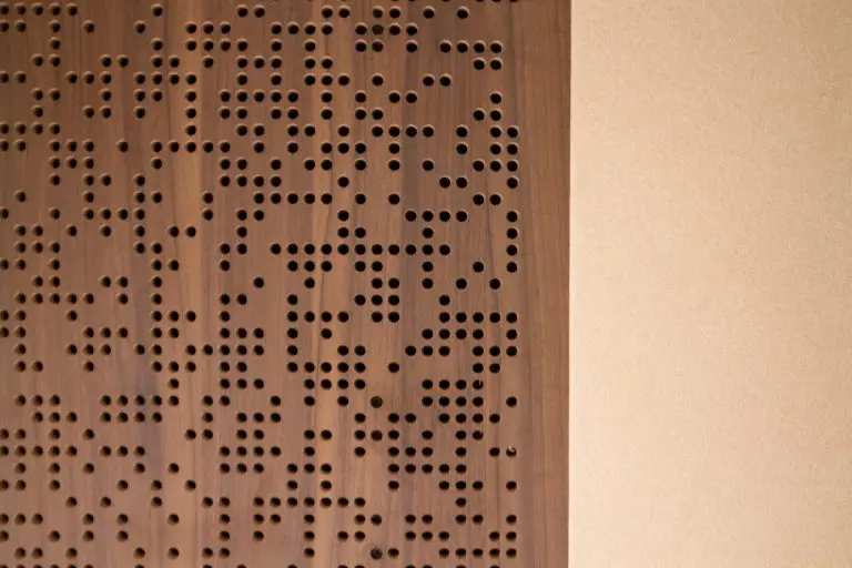 A close-up of a Timberix Acoustic Projects wooden panel with randomly spaced round holes on the left side, next to a plain beige wall on the right. The wood grain and perforations create a textured, modern design.