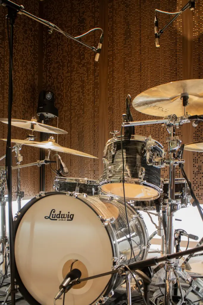 A Ludwig drum set with cymbals is set up on a stage, surrounded by several overhead microphones. The background features a brown, perforated Timberix Acoustic Projects panel.