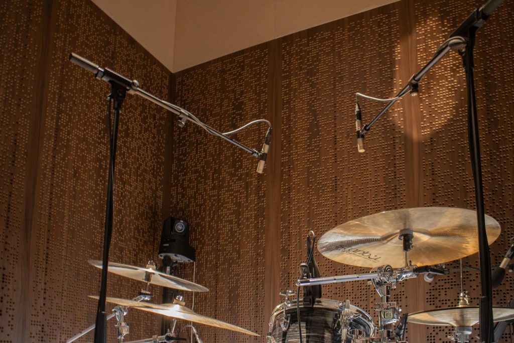 A drum set is positioned in a recording studio with Timberix Acoustic Projects wooden panels on the walls. Overhead condenser microphones are suspended above the drums, capturing the sound.