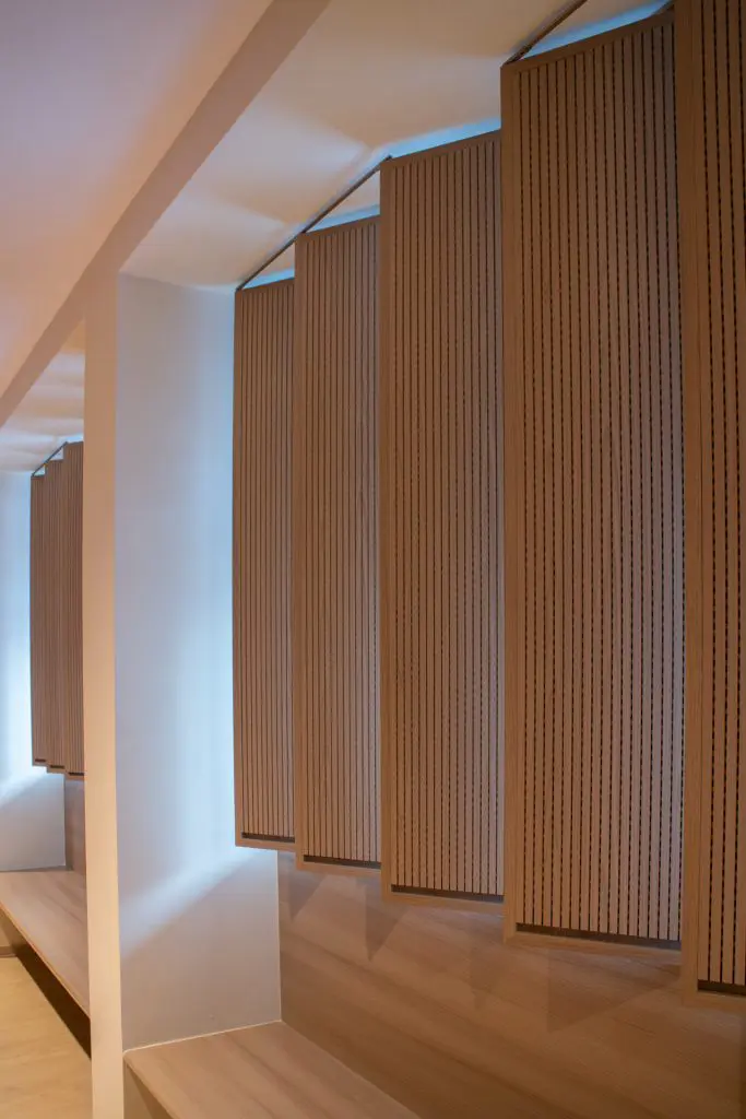 Vertical wooden panels with narrow perforations from Timberix Acoustic Projects hang from the ceiling along a corridor, creating a modern and minimalistic design. Soft lighting highlights the wood texture and brightens the light-colored walls and floor.