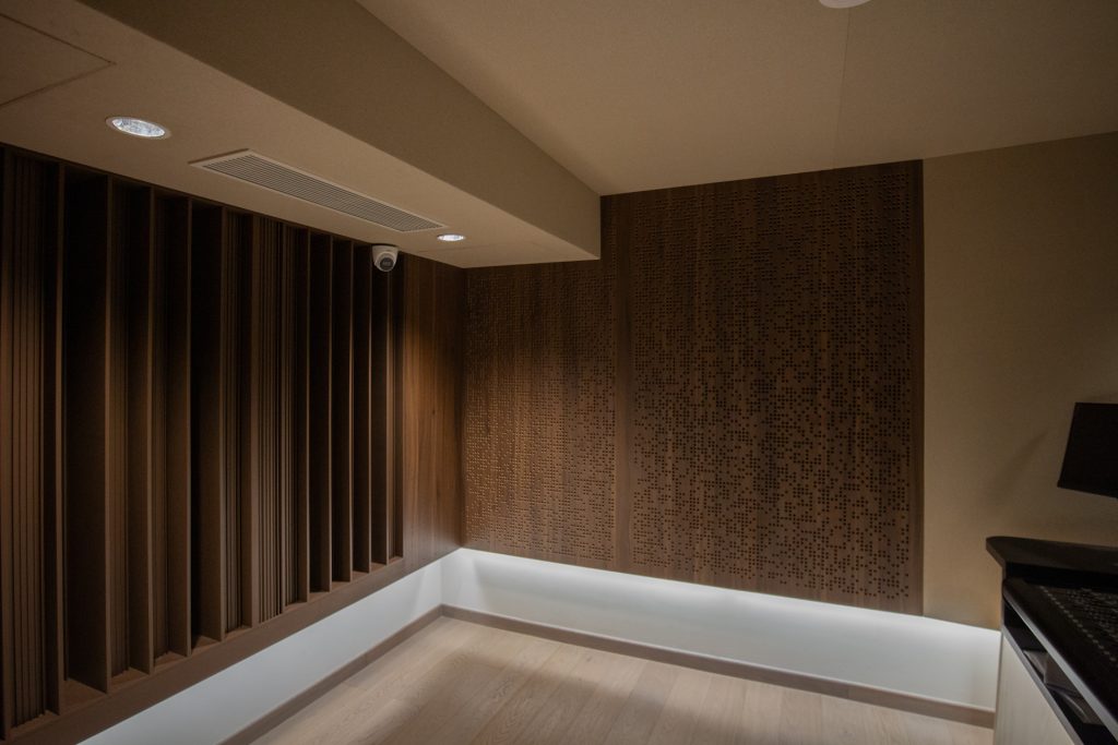 A modern room corner featuring Timberix Acoustic Projects: wooden acoustic panels on the walls—one with vertical slats, the other perforated—bathed in soft ceiling lights and complemented by a light wooden floor.