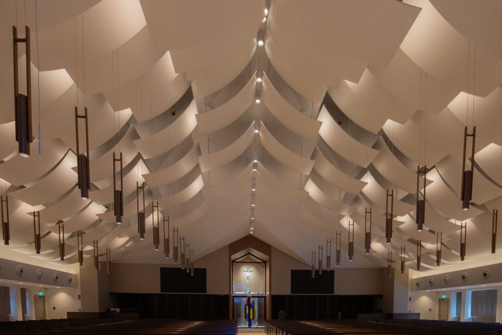 Interior of a modern church featuring Timberix Acoustic Projects, with dramatic geometric white ceiling panels, symmetrical hanging lights, empty pews, and a striking stained glass window above the entrance at the far end.