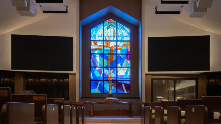 A church interior with Timberix Acoustic Projects enhancing the wooden pews, all facing a large, colorful stained glass window with abstract designs and a cross, flanked by two black rectangular screens on either side.