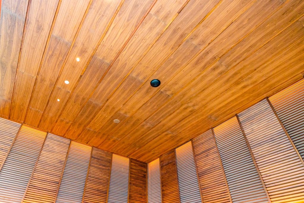 Angled view of a modern room’s wooden ceiling and upper walls, featuring narrow, horizontal wood panels and slats in warm tones—showcasing sustainable timber ceilings that meet green building codes, with recessed lights and a central black fixture.