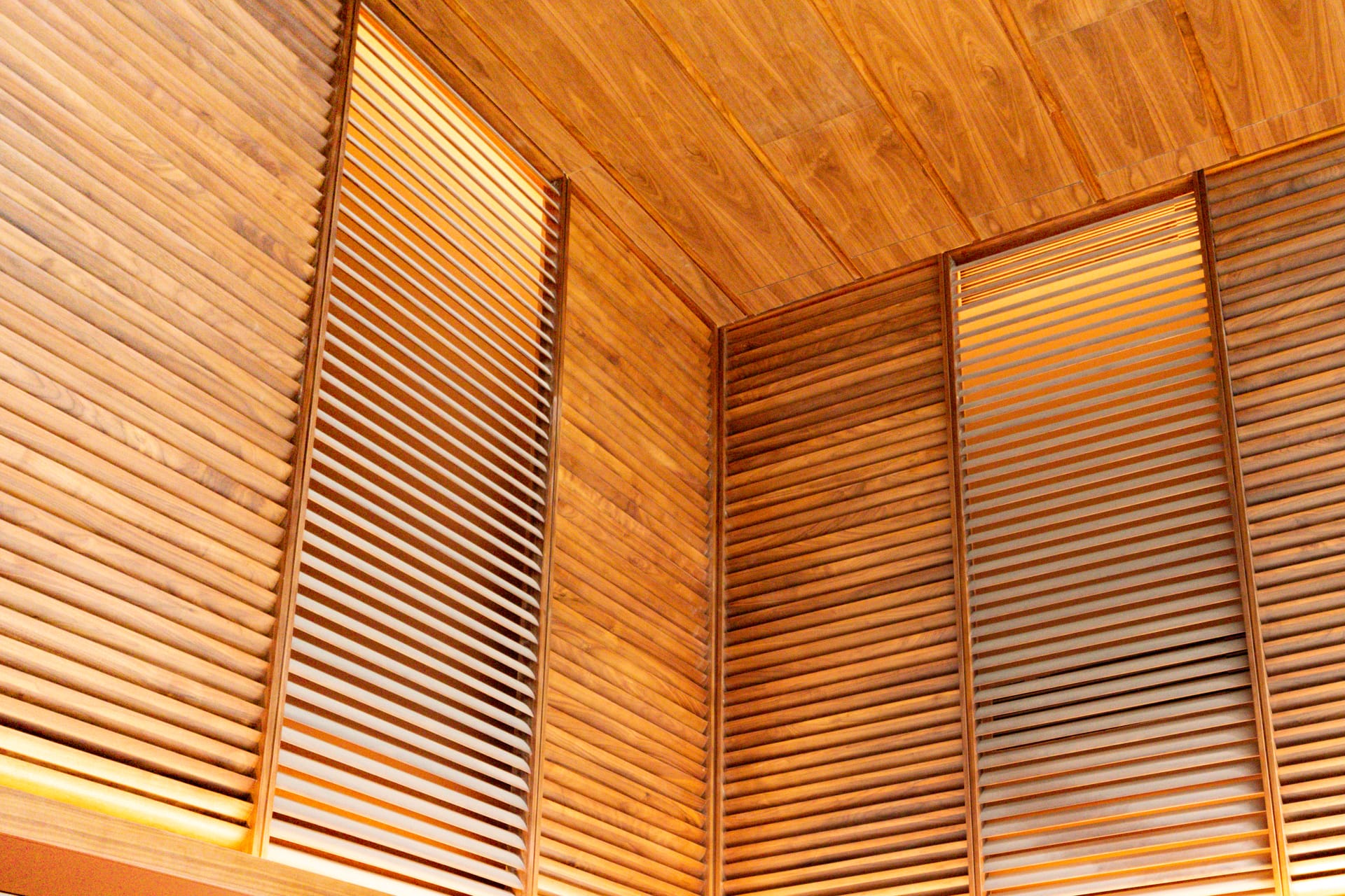 Wooden slatted wall panels and sustainable timber ceilings that meet green building codes create a warm, modern interior. Vertical and horizontal slats let light filter through, highlighting the wood’s natural grain and golden tones.