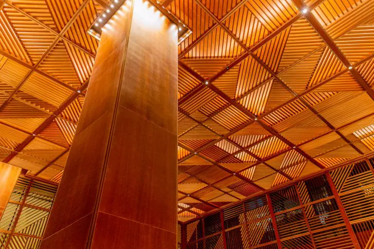 Geometric wooden ceiling with diagonal slats, creating a striking pattern. Sustainable timber ceilings that meet green building codes are complemented by a tall wooden column and warm lighting that highlights the intricate texture and matching lattice walls.