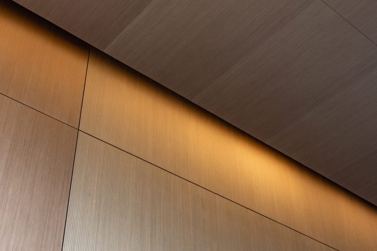 A modern interior wall and ceiling with light brown, multi-certified timber panels, intersecting lines, and warm indirect lighting creating a soft glow along the upper edge for high-performance interiors.
