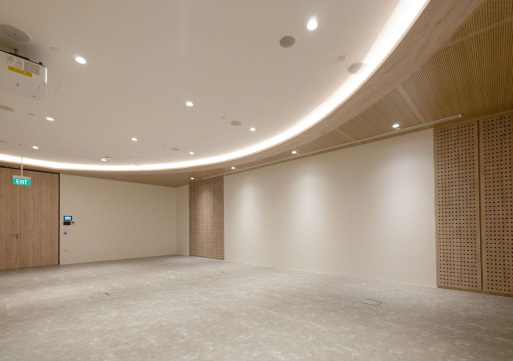 A modern, empty room with light-colored walls and flooring, wood panel accents, a curved ceiling with recessed lighting, a projector, and an illuminated exit sign above a closed wooden door.