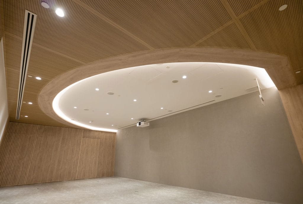 A modern, minimalist room with light wood paneled walls and a large oval-shaped recessed ceiling light. The space is empty, with a gray floor and a projector mounted on the ceiling.