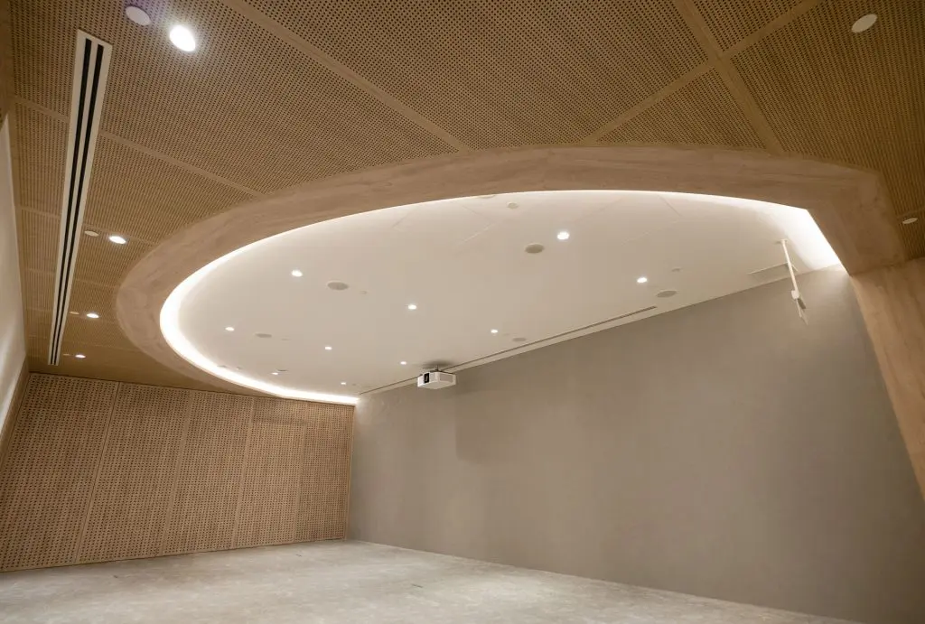 A modern, minimalist room with light wood paneled walls and a large oval-shaped recessed ceiling light. The space is empty, with a gray floor and a projector mounted on the ceiling.