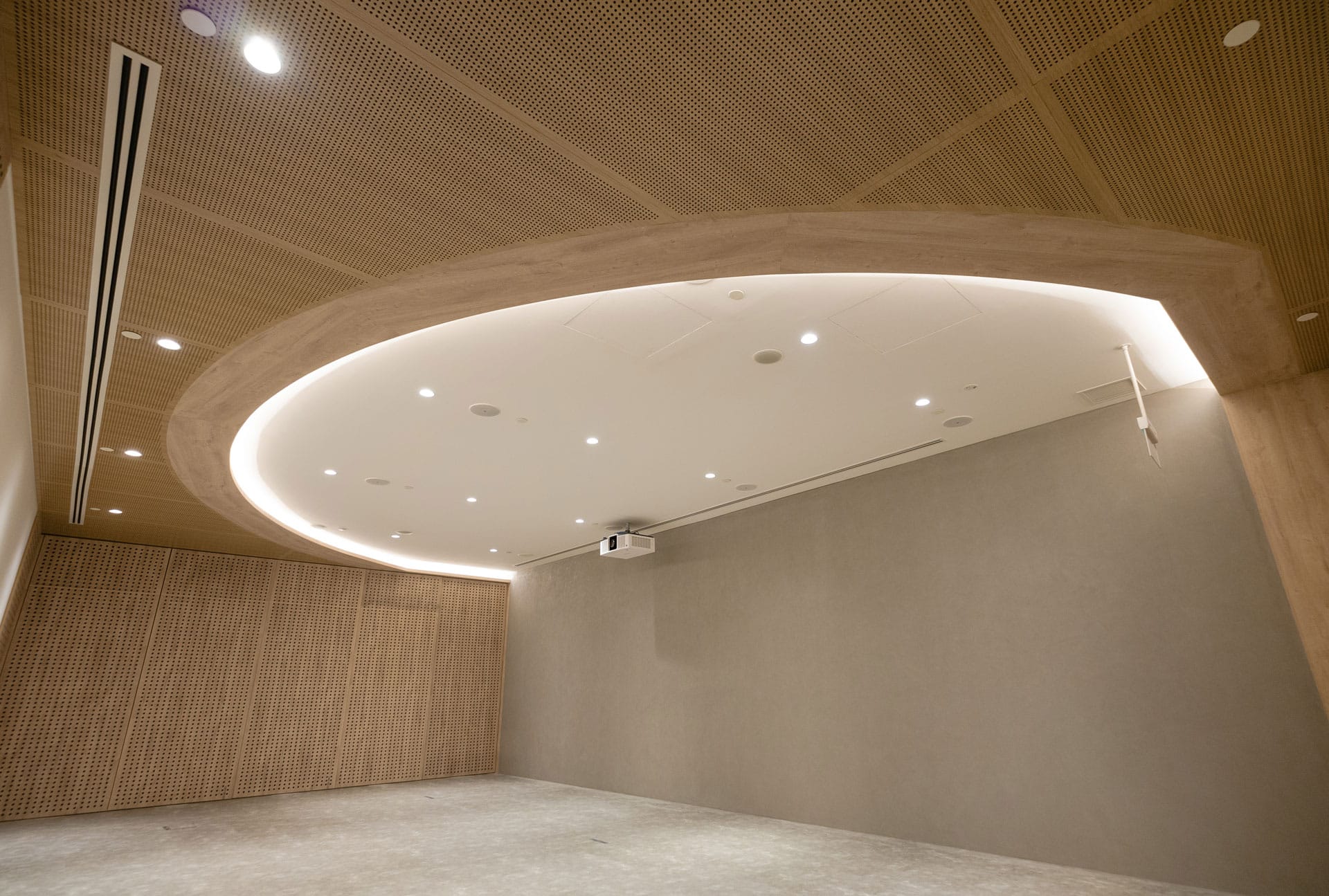 A modern, minimalist room with light wood paneled walls and a large oval-shaped recessed ceiling light. The space is empty, with a gray floor and a projector mounted on the ceiling.
