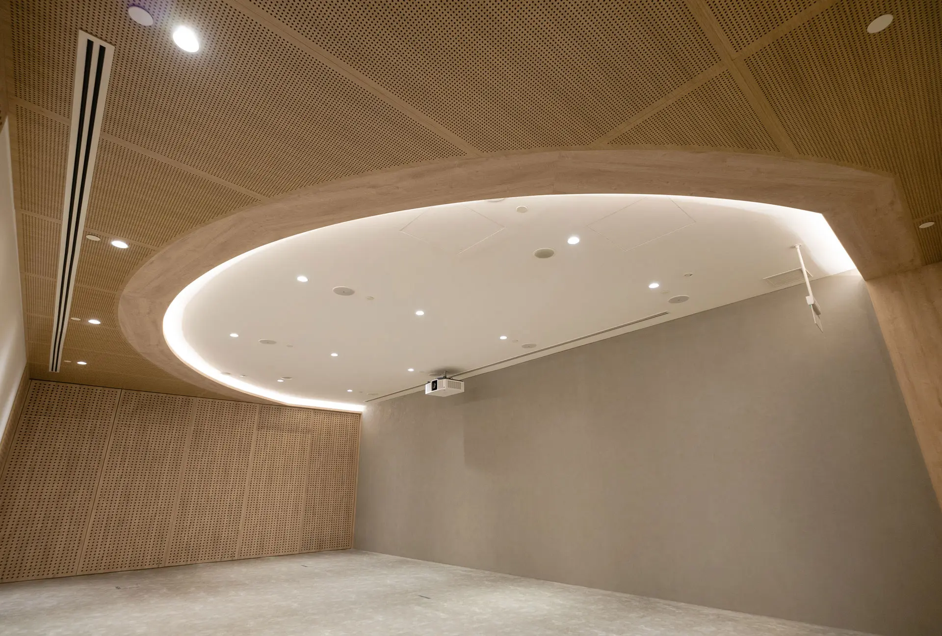 A modern, minimalist room with light wood paneled walls and a large oval-shaped recessed ceiling light. The space is empty, with a gray floor and a projector mounted on the ceiling.