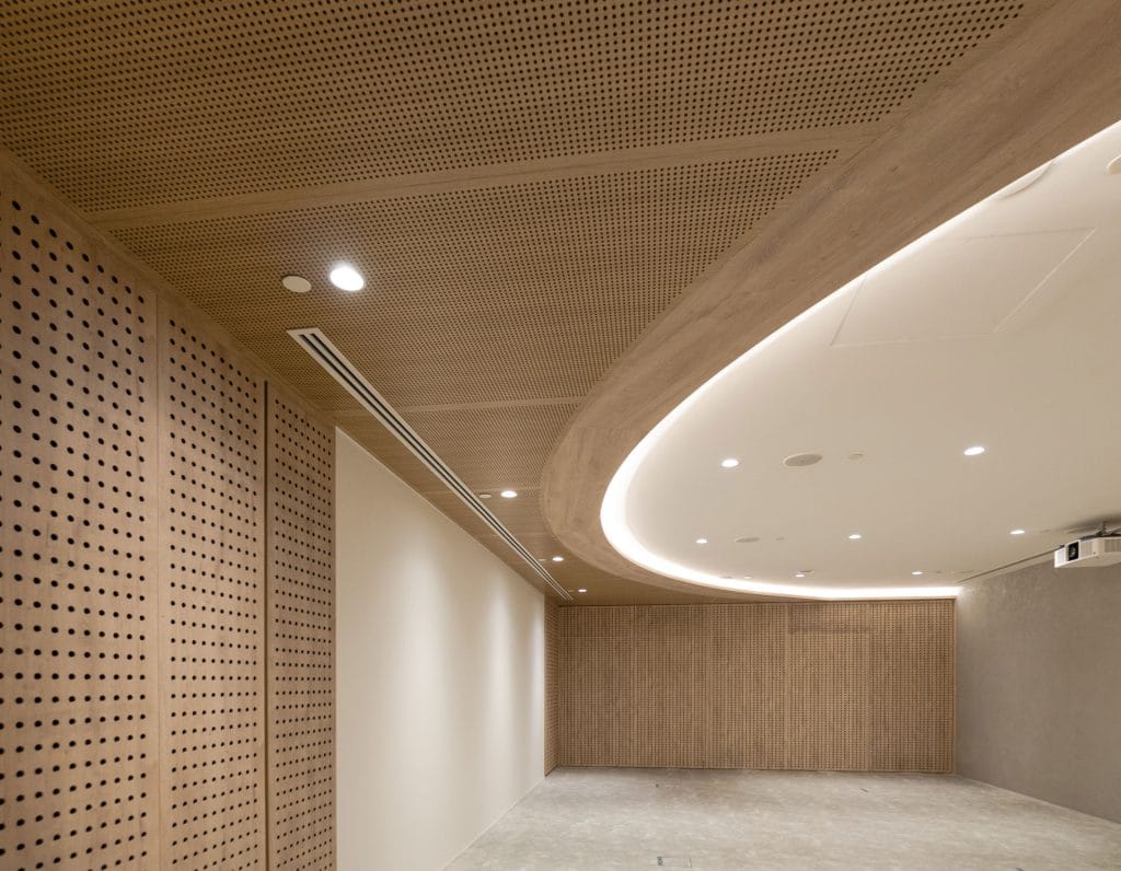 A modern, empty room with beige perforated wood panel walls and ceiling, recessed lighting, and a curved, illuminated ceiling feature. The floor appears unfinished, and the overall space looks clean and minimalistic.