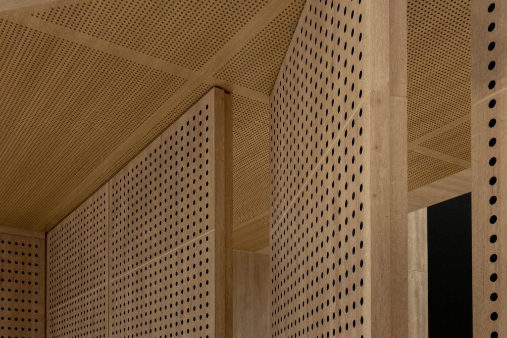 Geometric wooden panels with a perforated pattern of evenly spaced circular holes, forming walls and ceiling in an architectural interior. The wood has a light, natural finish.