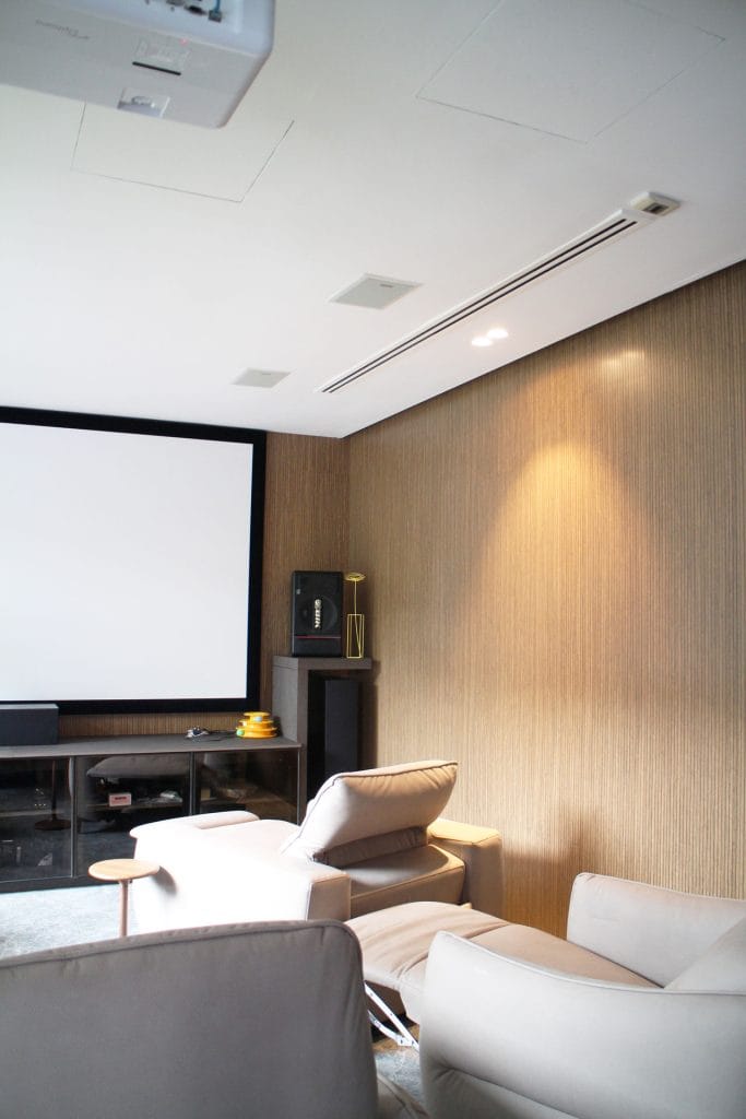 A modern home theater room with beige recliner chairs, a large projection screen, a wooden accent wall, shelves with audio equipment, and soft overhead lighting.