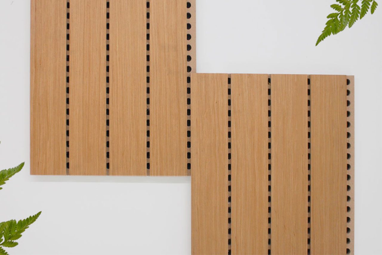 Two Timberix Grooved Acoustic Panels in light wood with vertical black grooves are mounted on a white wall, slightly offset from each other. Green fern leaves partially frame the image from the corners.