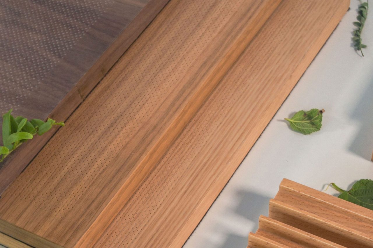 Close-up of micro perforated wood panels with finely drilled holes, arranged on a light surface and accented with a few green leaves. The panels display varied wood tones and distinct grain patterns.