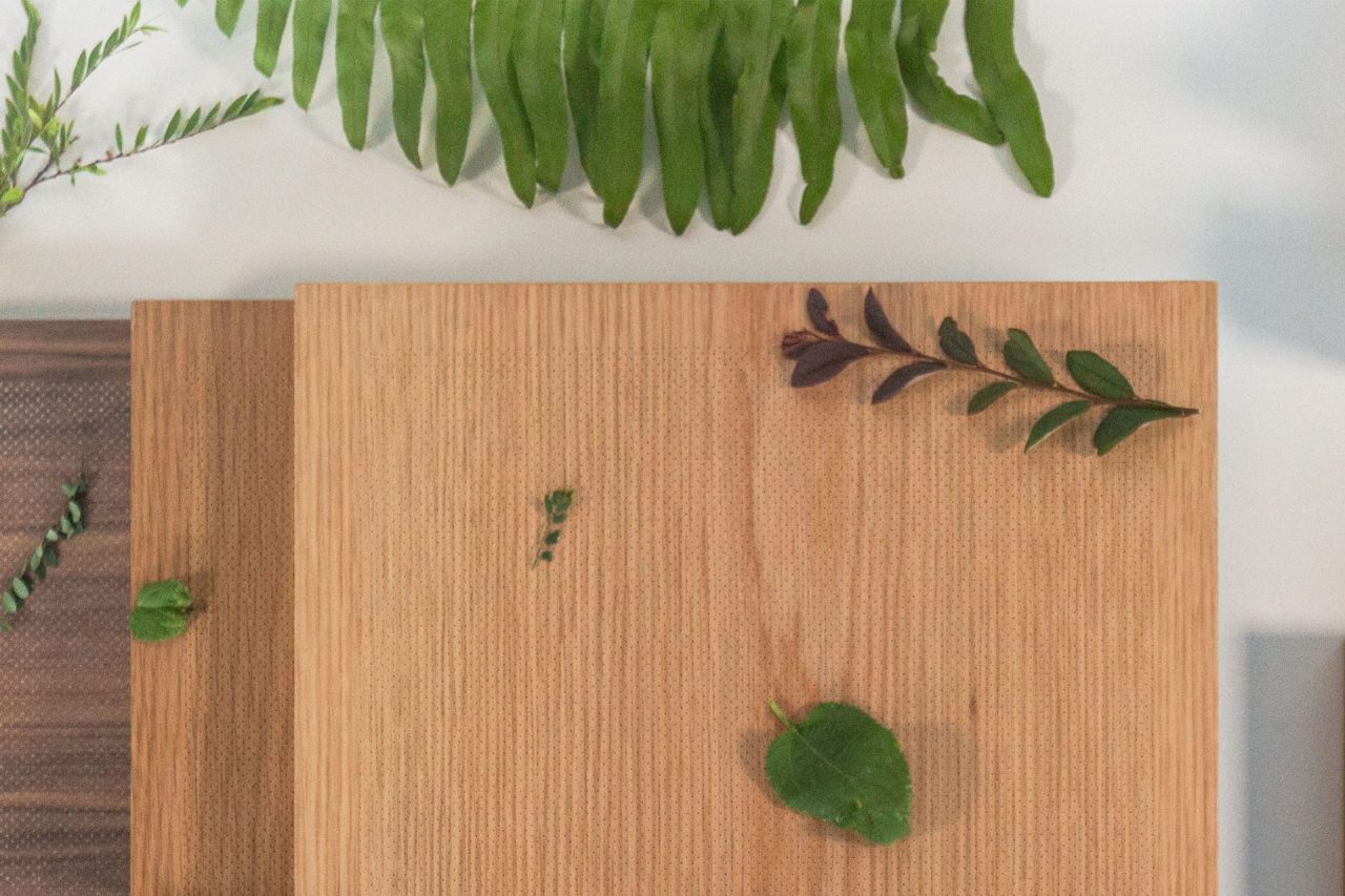Two wooden cutting boards, one stacked partly over the other, with small green leaves and sprigs on them. A large fern leaf and other greenery are placed at the top of the image on a white surface.