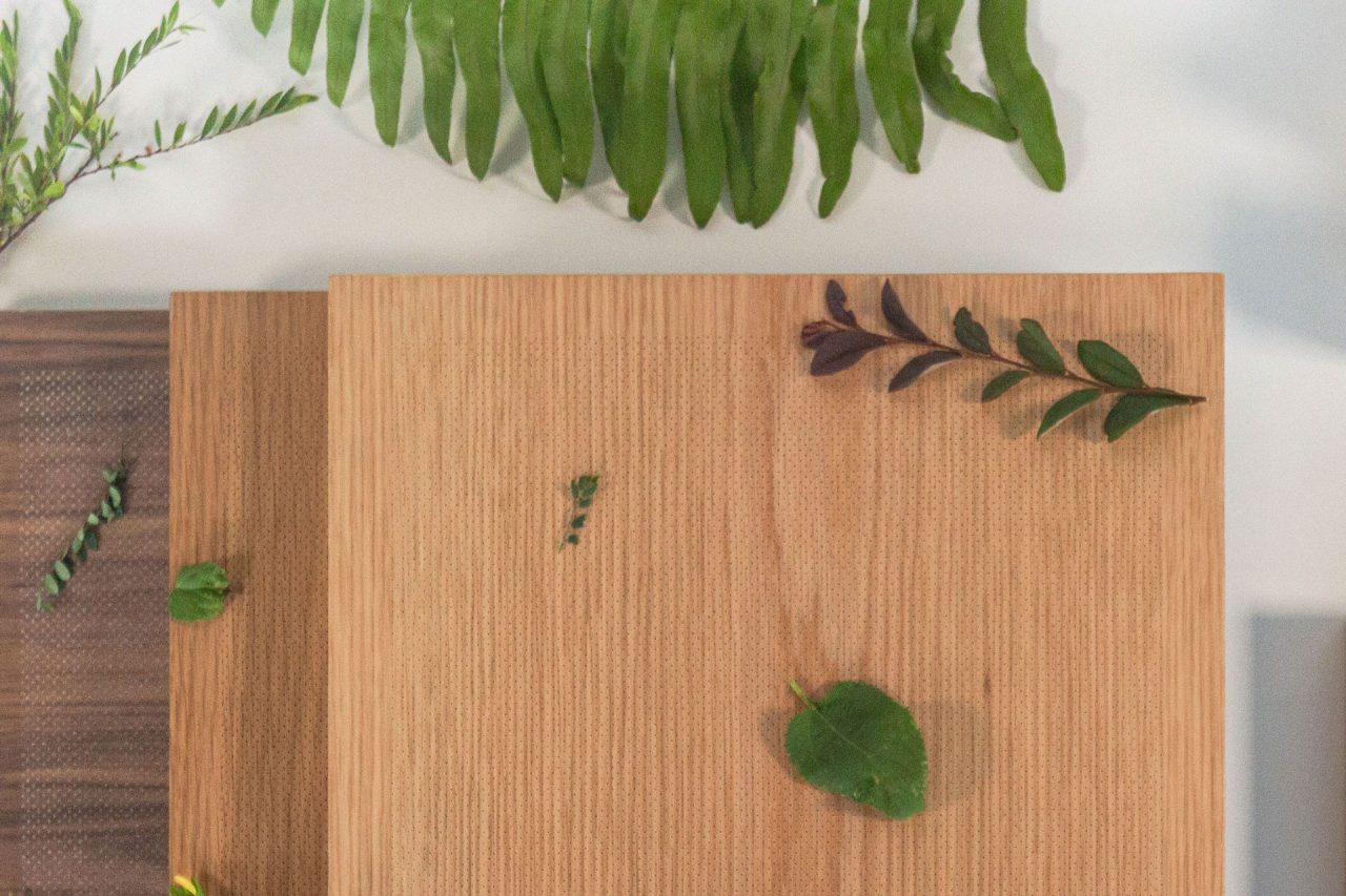 A wooden cutting board with a few green leaves and sprigs arranged on top, alongside another board and fern leaves at the top of the image. The background is light and uncluttered, evoking the clean look of micro perforated wood panels.
